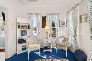 Ein Zimmer mit zwei Korbstühlen, einem weißen Regal mit Büchern und einer Lampe sowie einem Fenster mit Vorhängen.