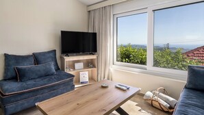 A modern living room with a sofa, a coffee table, a TV, and a large window offering a view of greenery and buildings. - 3-bedroom eco villa in excellent Dilinata, family / friends friendly (Dilinata)