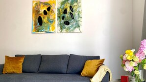 Um sofá cinzento com duas almofadas mostarda, uma pequena mesa de apoio branca com um vaso de flores, e duas pinturas abstratas na parede.