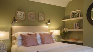 Una cama bien hecha con almohadas, una lámpara de noche e impresiones botánicas enmarcadas en la pared.