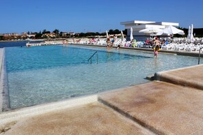 En swimmingpool med klart vand, omgivet af liggestole og parasoller, med en bygning i baggrunden.
