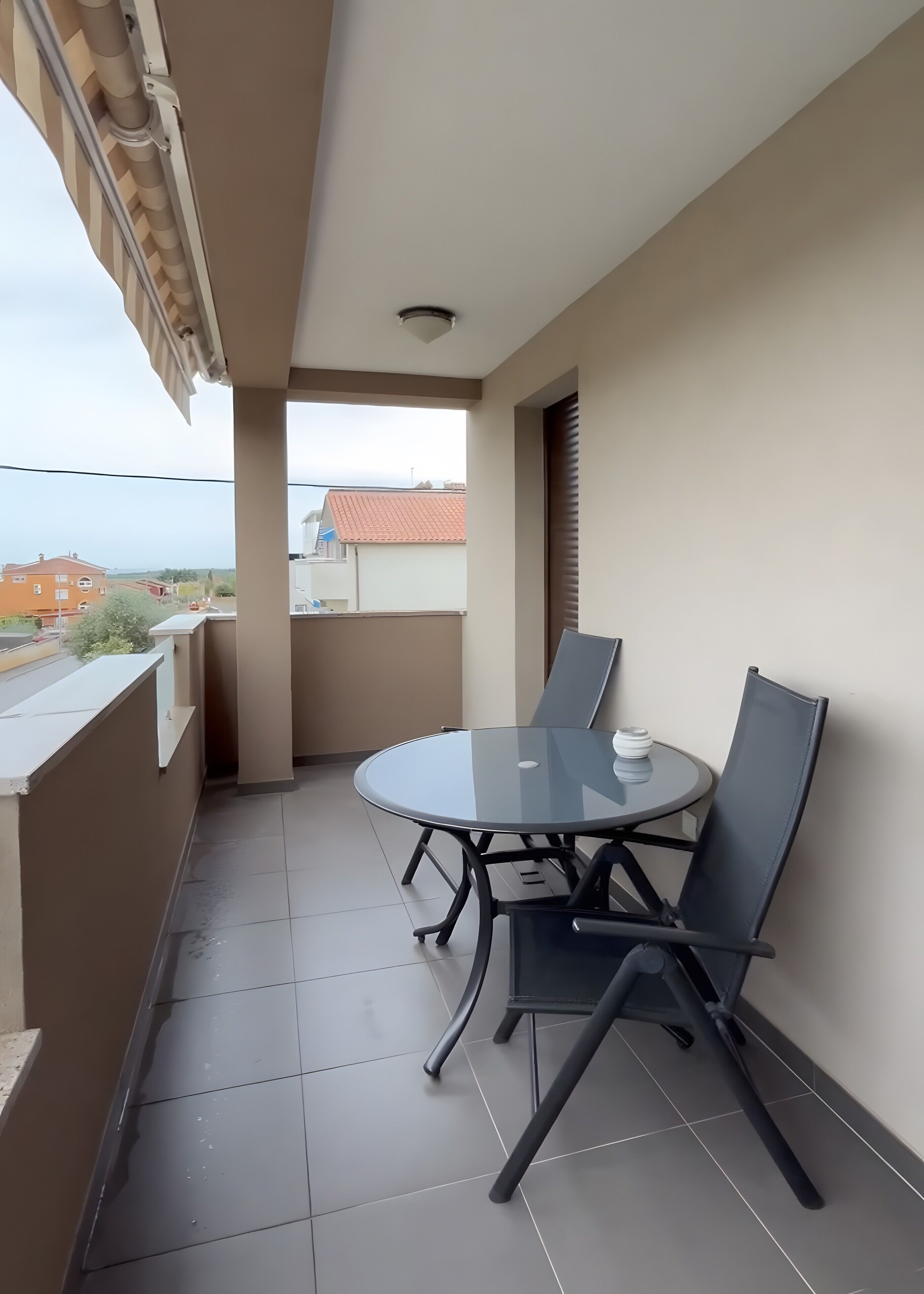 A balcony with a round glass table and two chairs, a view of the sea, and a tiled floor.