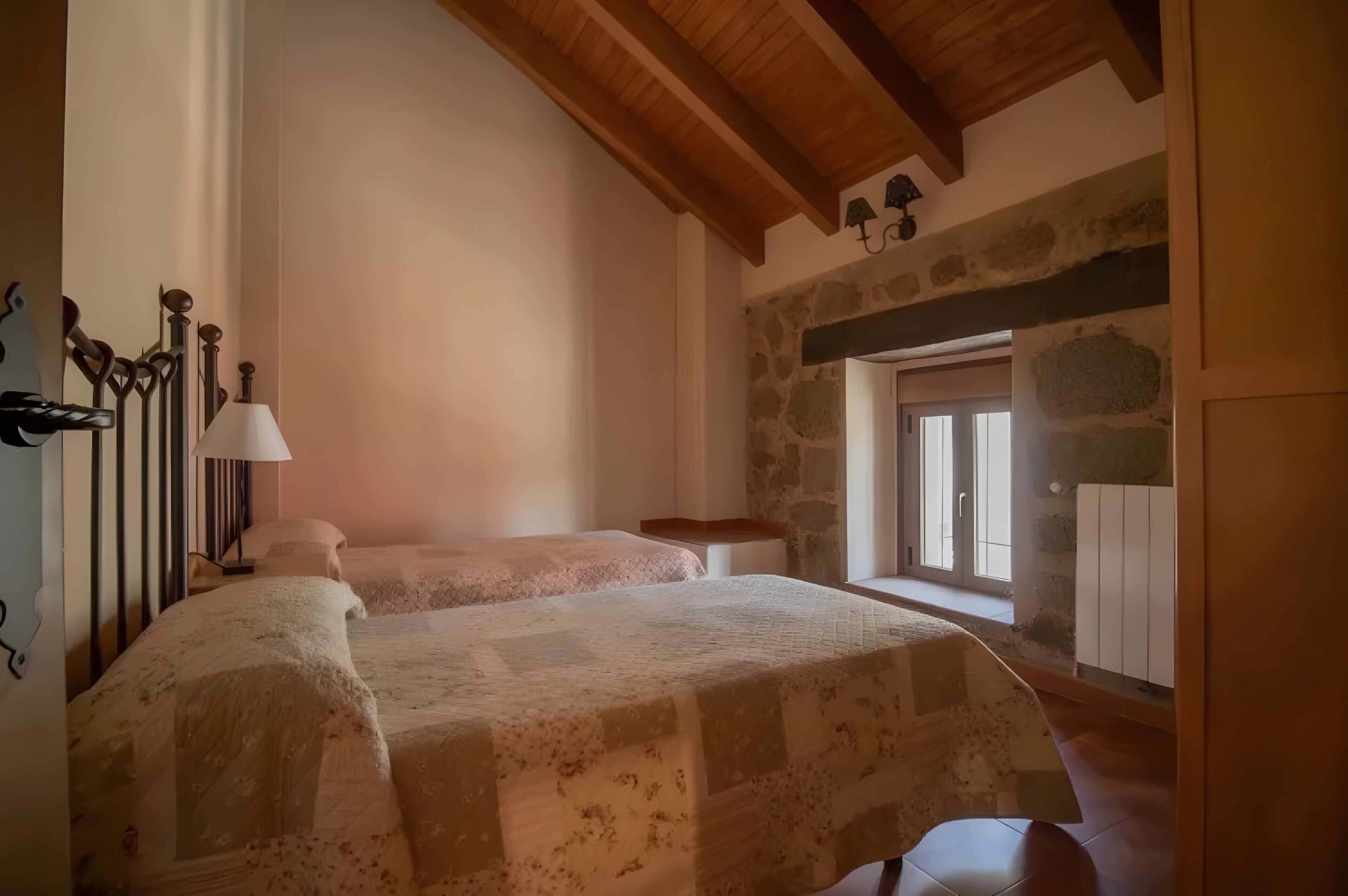 A bedroom with two beds, a stone wall, a wooden ceiling, and a window.