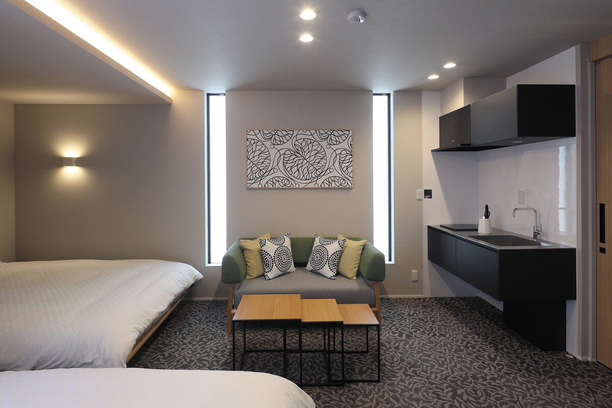 Une chambre d’hôtel avec deux lits, un canapé agrémenté de coussins à motifs, une petite table en bois, une kitchenette avec un évier et une œuvre d’art accrochée au mur.