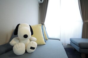 Un juguete de perro suave en un sofá con un cojín azul claro, una almohada amarilla y un sofá gris en una habitación con ventana y cortinas.