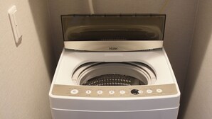 A compact, white, front-loading washing machine with a transparent door, a control panel with various buttons, and a power cord plugged into a wall socket. - Koti Sopo Universal Bay 2 by Liaison (Osaka)