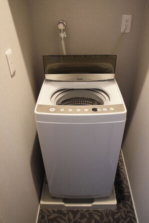 Une machine à laver compacte, blanche, à chargement frontal, dotée d’une porte transparente, d’un panneau de commande avec divers boutons et d’un câble d’alimentation branché sur une prise de courant.