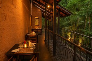 Un área de comedor al aire libre con mesas y sillas, un paseo techado y vistas a abundante vegetación.