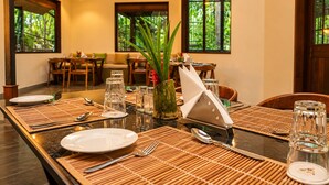 A dining area with a long table set for two, surrounded by chairs, with a view of greenery through large windows.