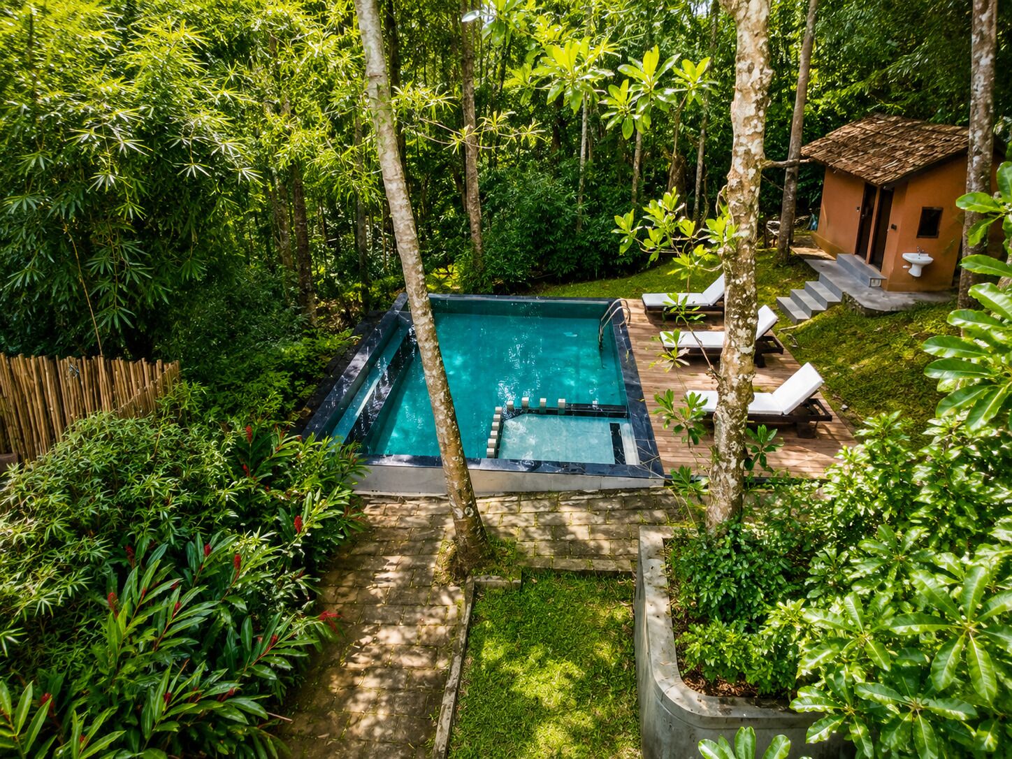 Una piscina rectangular con cubierta de madera, rodeada de abundante vegetación y un pequeño edificio con tejado de baldosas.
