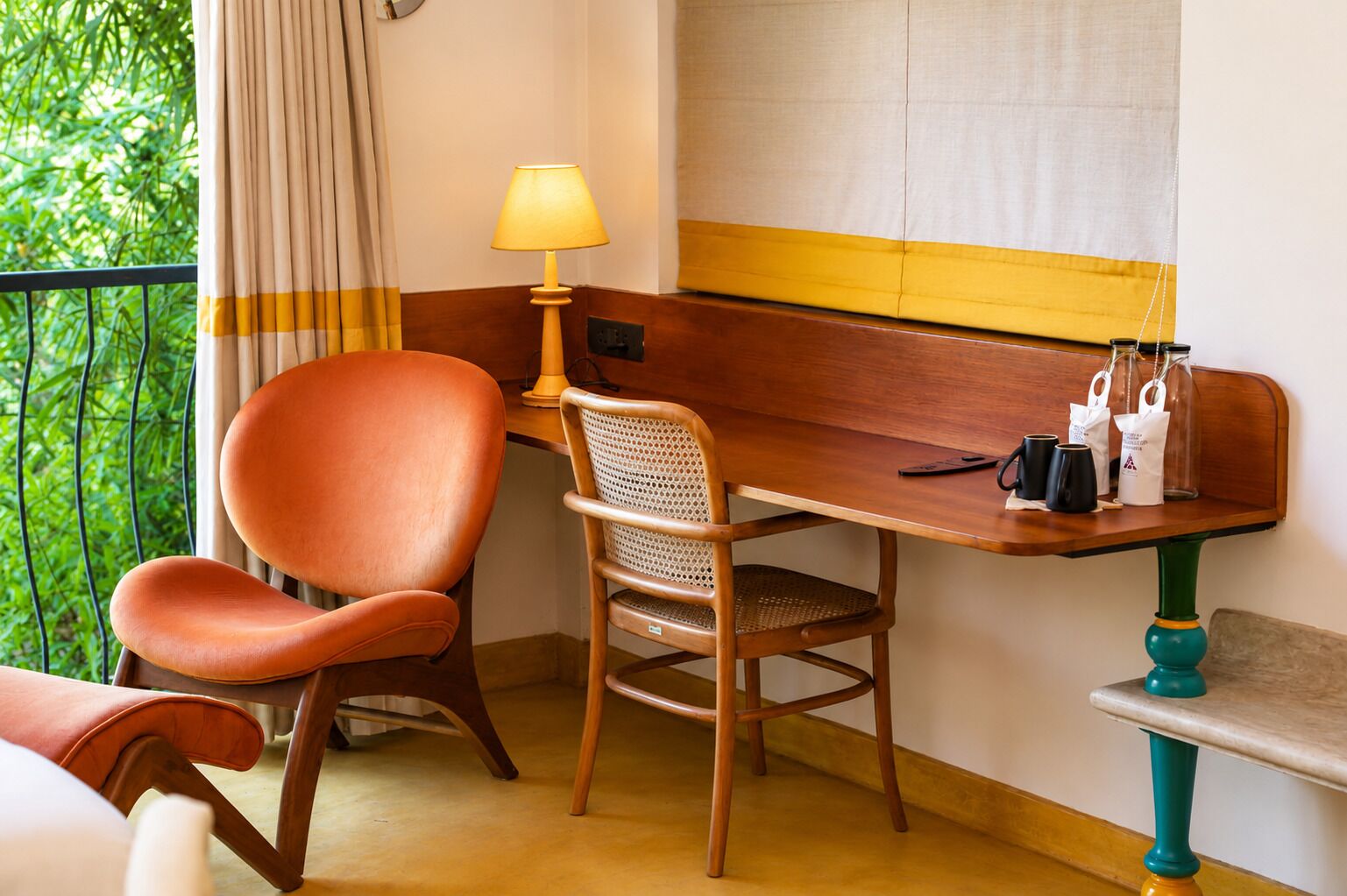 A room with a wooden desk, a chair, a lamp, and a view of greenery outside.