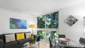 A modern living room with a sofa, coffee table, and chairs, large windows with a view of greenery, and a painting on the wall. - Modern 2BR Grove Condo w/ King & Balcony (Miami)
