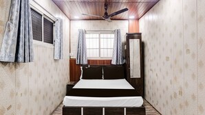 A bedroom with a bed, a ceiling fan, a window with curtains, and a wooden wardrobe. - Deluxe Hotel Garden Inn (Ambernath)