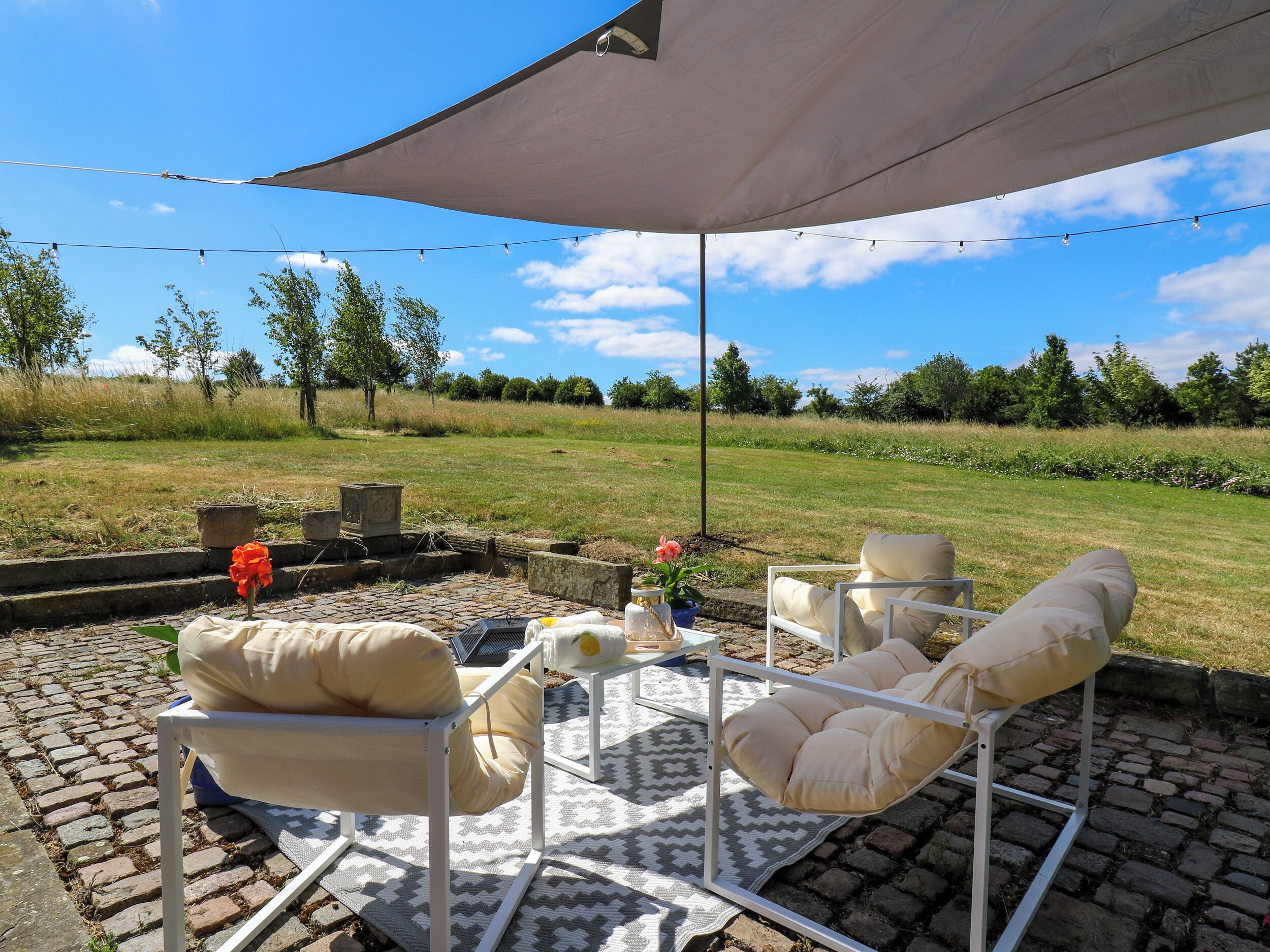 An outdoor patio with a white metal table, two white cushioned chairs, a grey patterned rug, and a large canopy.