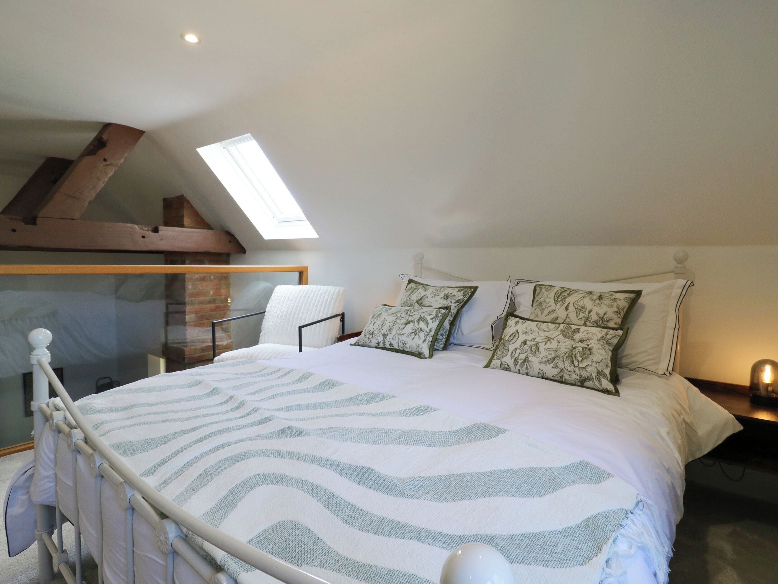 A bedroom with a sloped ceiling, a skylight, a bed with patterned pillows, a chair, and a bedside table.