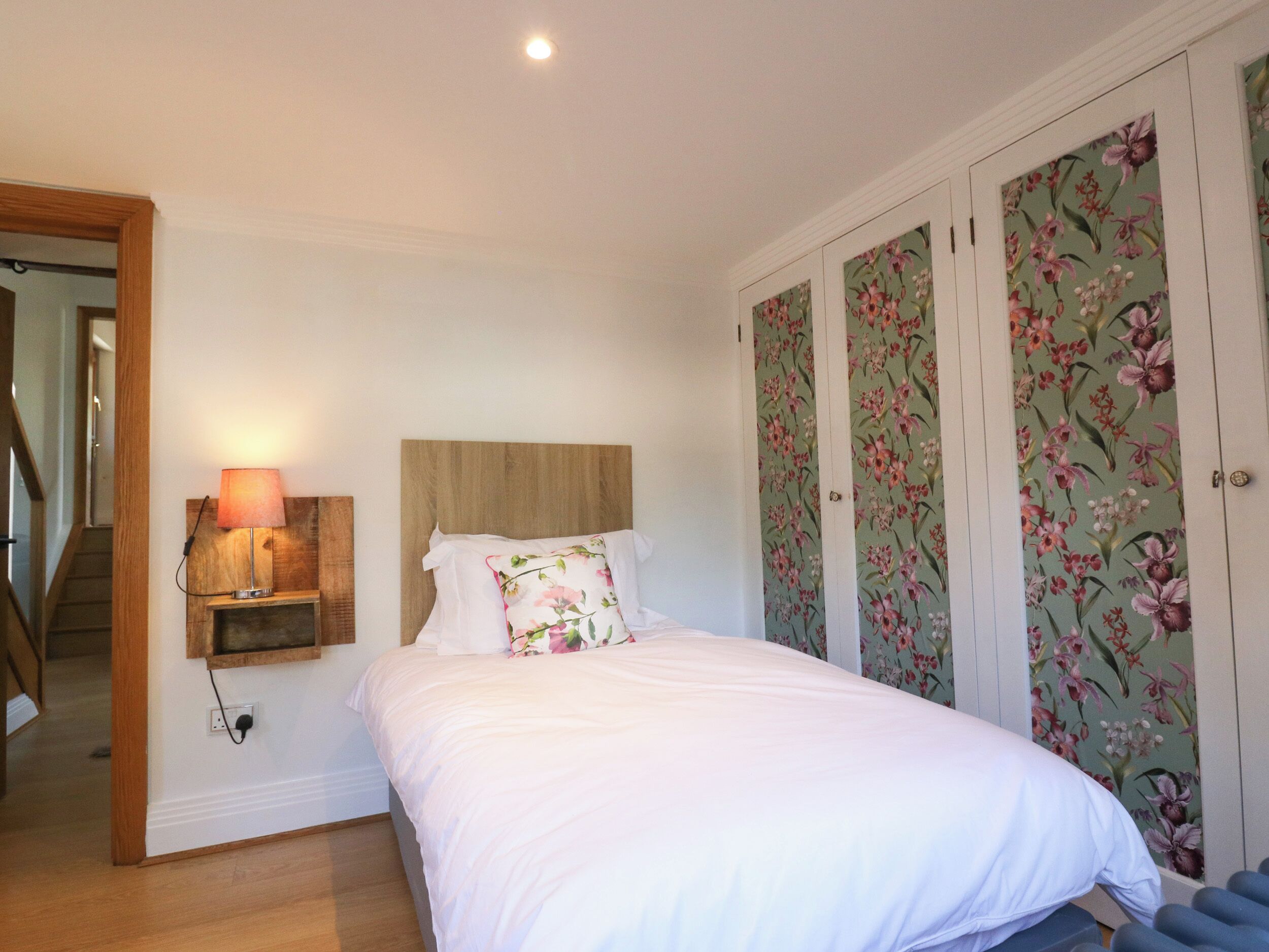 A bedroom with a bed, a nightstand with a lamp, a wooden headboard, and floral wallpaper.