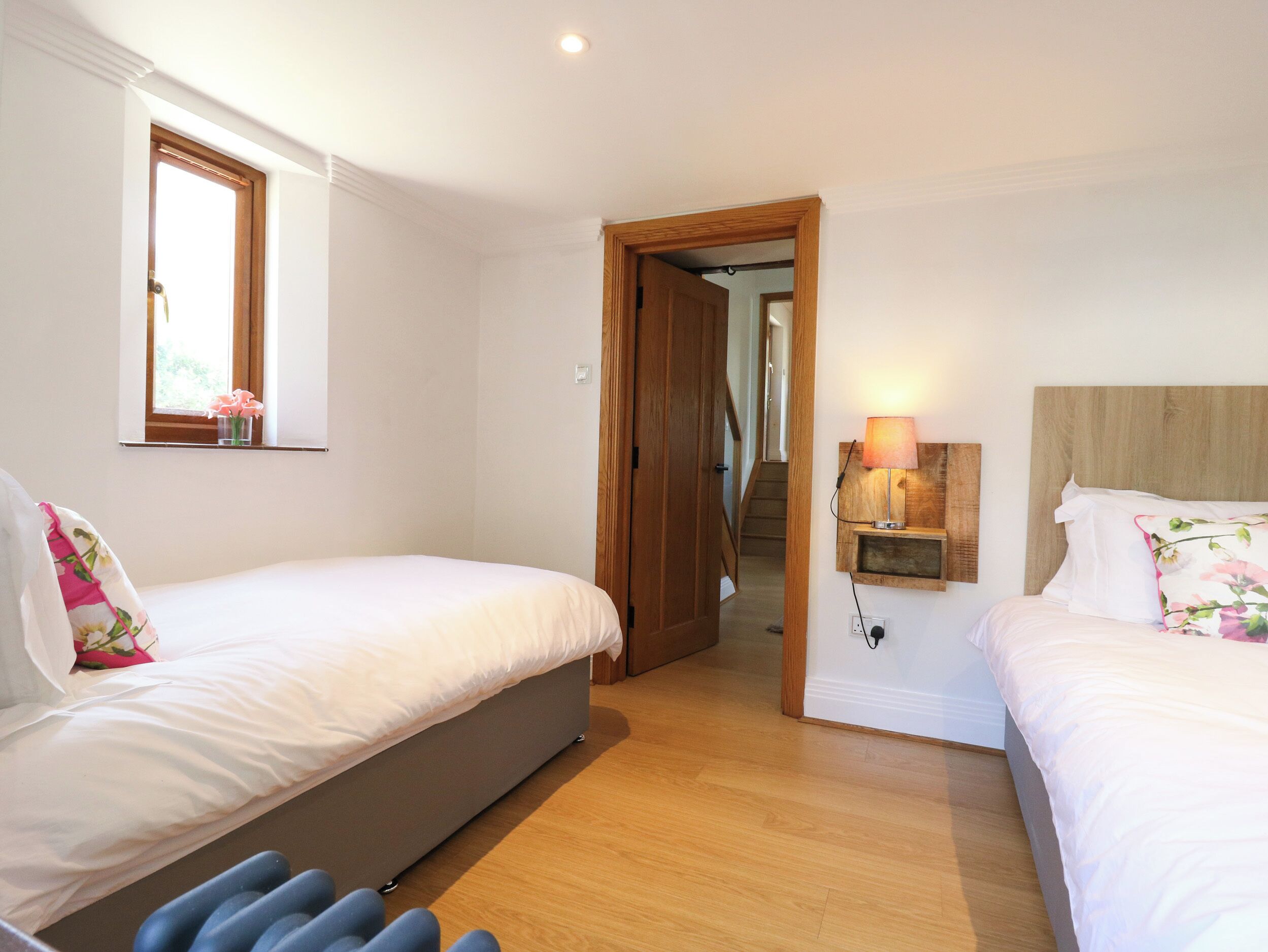 A bedroom with two beds, a wooden floor, a window with flowers, and a lamp on the wall.