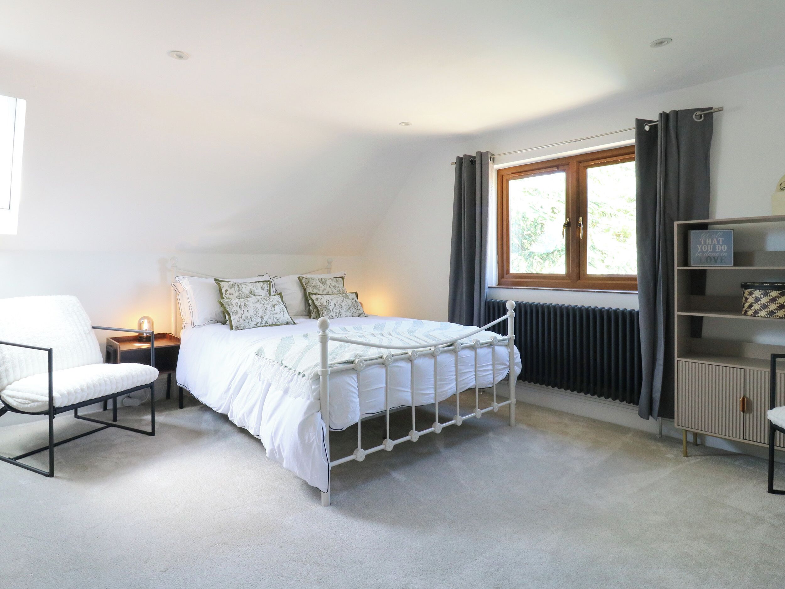 A bedroom with a white metal bed, a chair, a bedside table, a radiator, and a window with curtains.