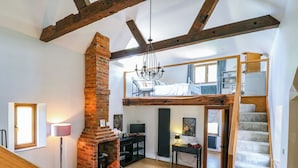 A loft-style living space with exposed wooden beams, a brick fireplace, a sofa, and a staircase leading to an upper level.