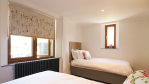 A bedroom with two beds, a radiator, a window with a floral curtain, and a small window above the bed.