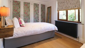 Un dormitorio con una cama, una mesita de noche, una lámpara, un radiador y una ventana con cortinas florales.