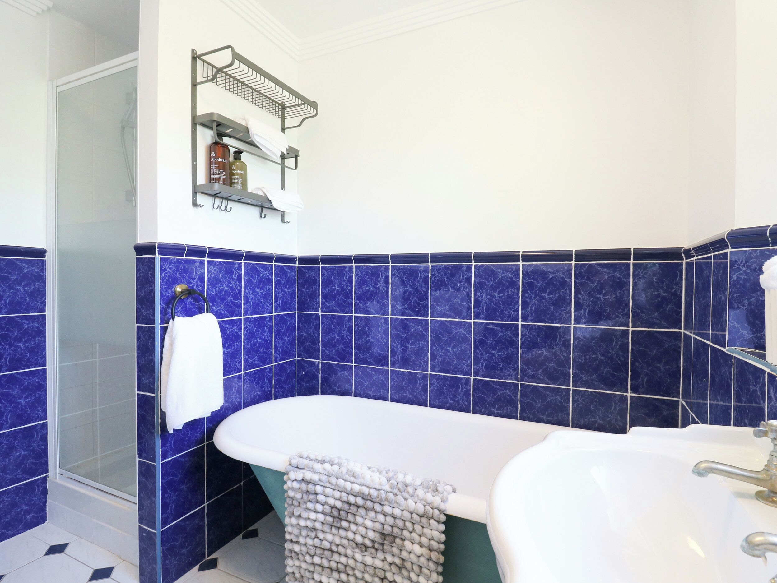 A bathroom with a bathtub, a shower, and a sink. There is a towel rack with a towel and a bottle of shampoo. The walls are tiled in blue.