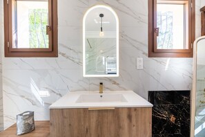 A bathroom with a marble wall, a wooden vanity, a white sink, a mirror with a light, and a black marble fireplace.