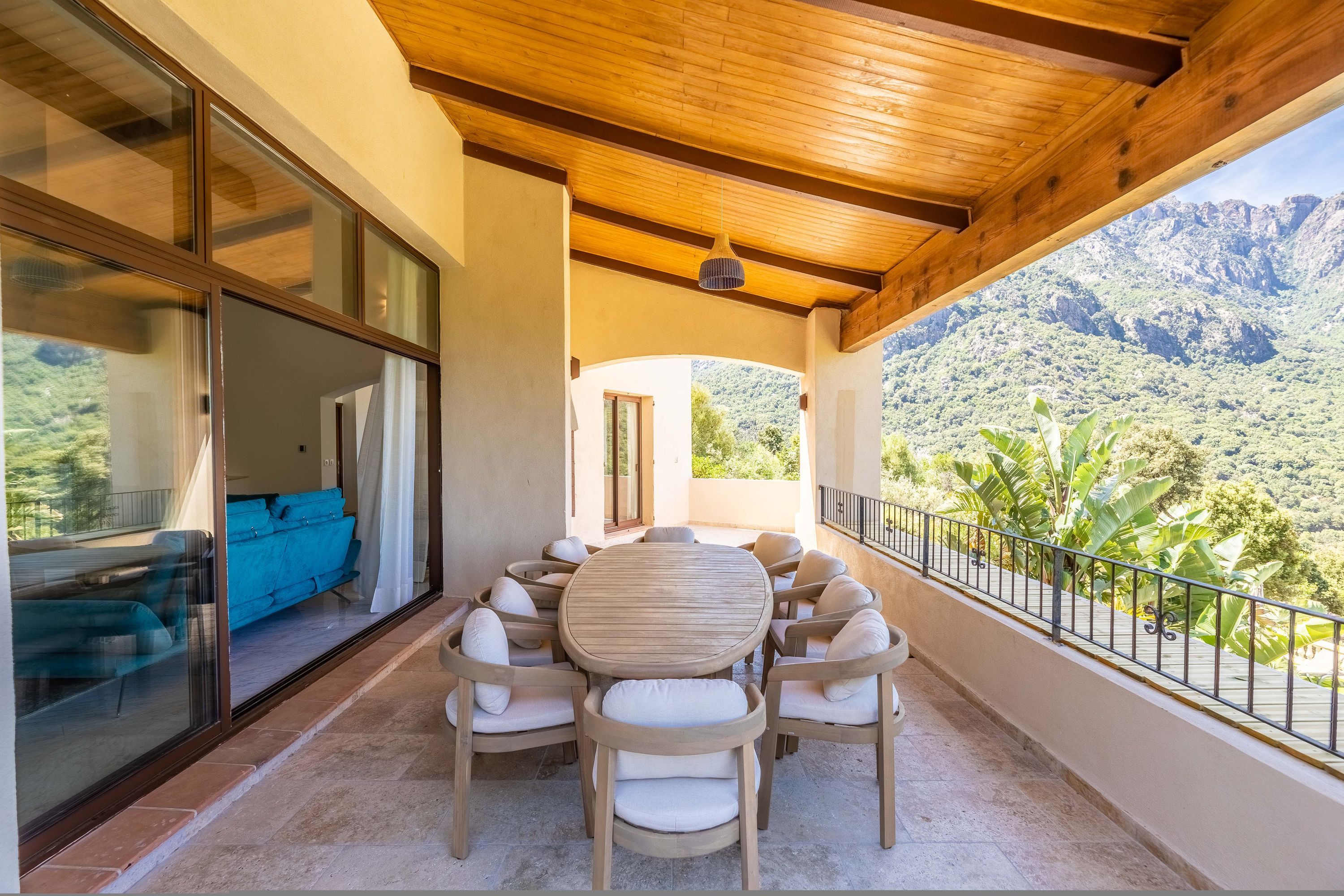A covered outdoor seating area with a wooden table and chairs, a sliding glass door leading to a room, and a view of mountains.