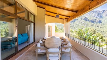 A covered outdoor seating area with a wooden table and chairs, a sliding glass door leading to a room, and a view of mountains.