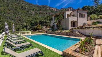 A swimming pool with lounge chairs, a stone building, and a mountainous backdrop.