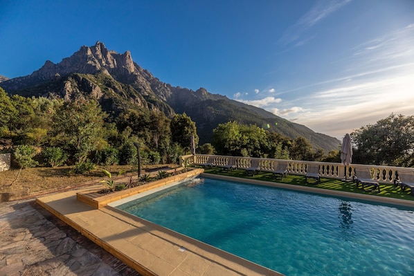 A pool with a mountain backdrop, a stone deck, and a pergola with umbrellas.