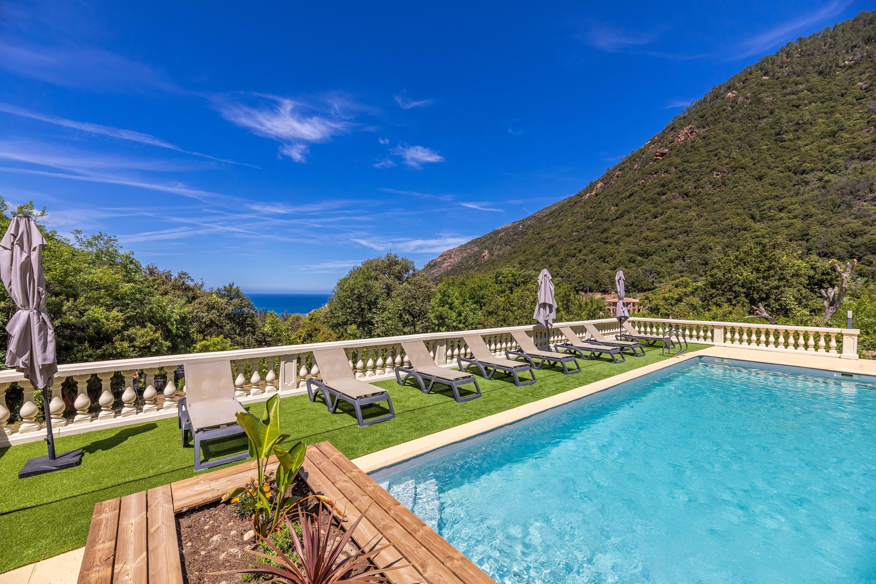 Ein Swimmingpool mit Liegestühlen und Sonnenschirmen, umgeben von einer weißen Balustrade, mit Blick auf einen grünen Hügel und das Meer.