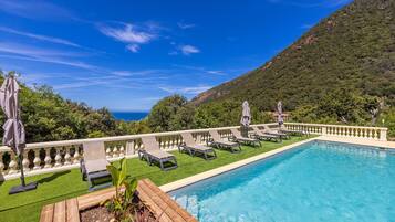 Ein Swimmingpool mit Liegestühlen und Sonnenschirmen, umgeben von einer weißen Balustrade, mit Blick auf einen grünen Hügel und das Meer.