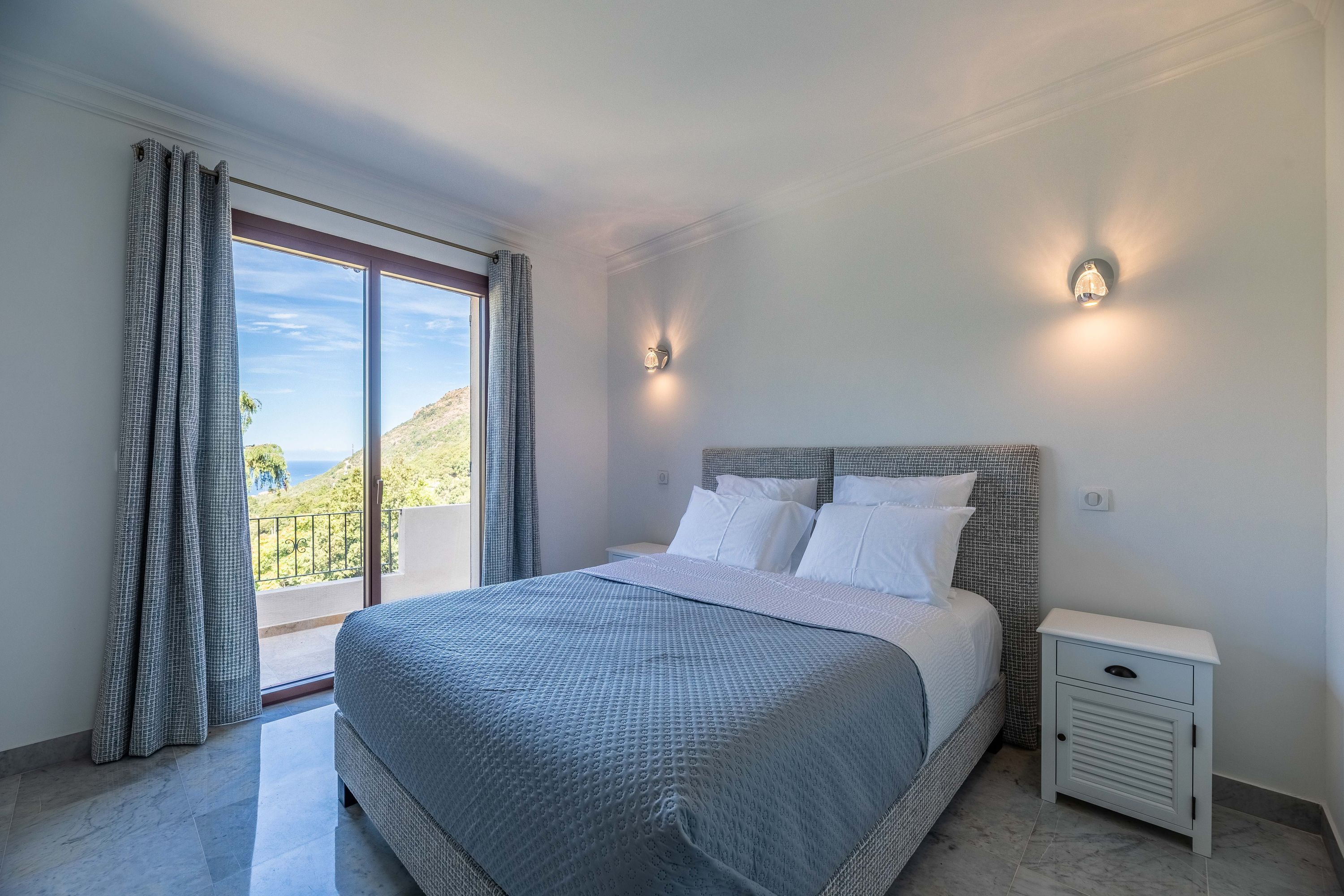 A bedroom with a large bed, a nightstand, two wall sconces, and a view of the outdoors.