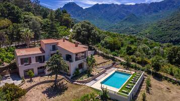 A two-story house with a swimming pool, surrounded by lush greenery and mountains.