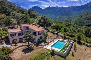 Una casa de dues plantes amb una piscina, envoltada de vegetació exuberant i muntanyes.