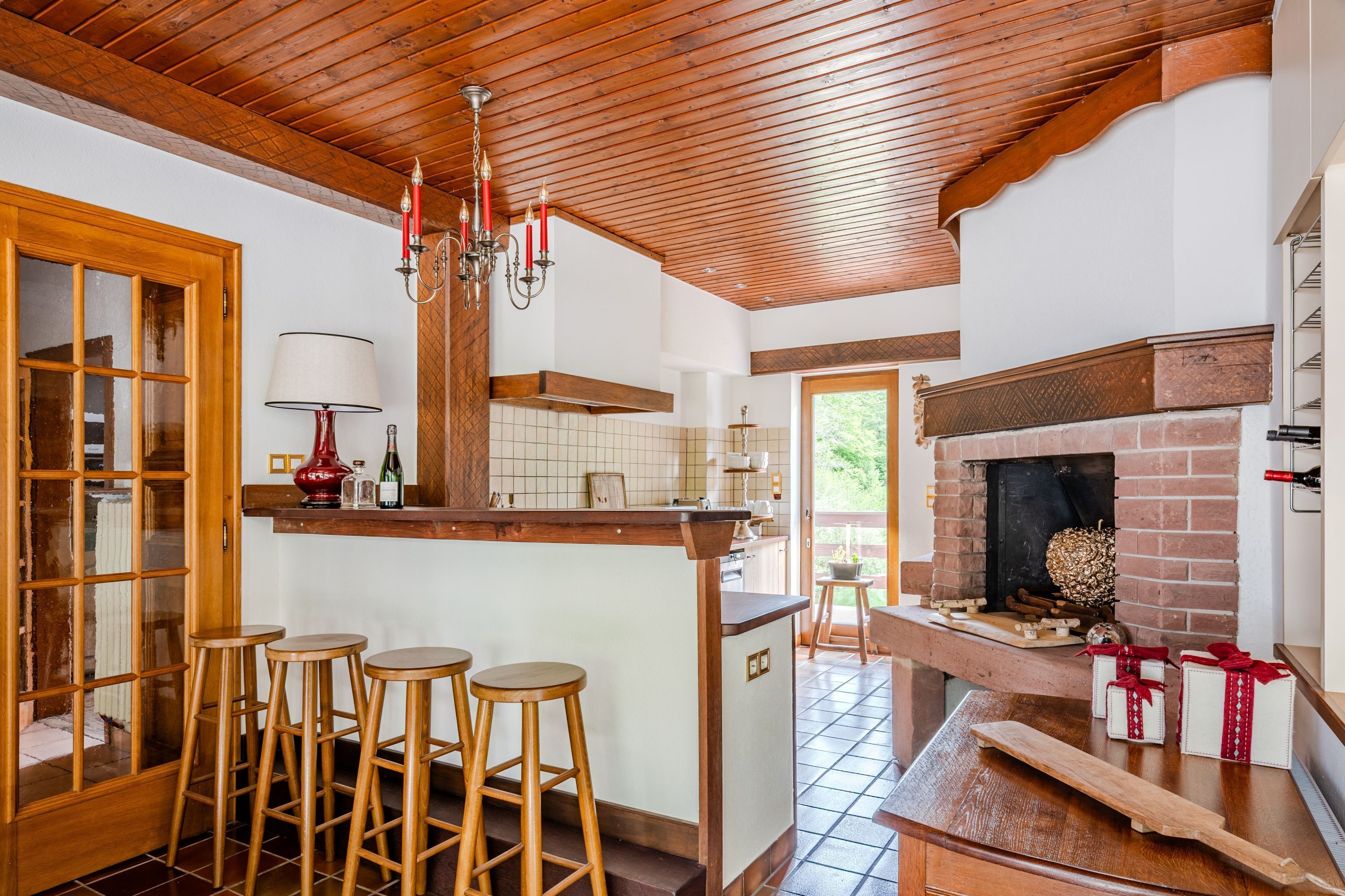 Une cuisine avec des poutres en bois, un coin bar avec des tabourets, une cheminée et une vue sur l’extérieur.