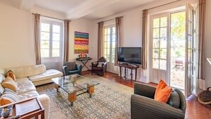 Ein Wohnzimmer mit einer Couch, Sesseln, einem Couchtisch, einem Fernseher und einem großen Fenster mit Vorhängen.