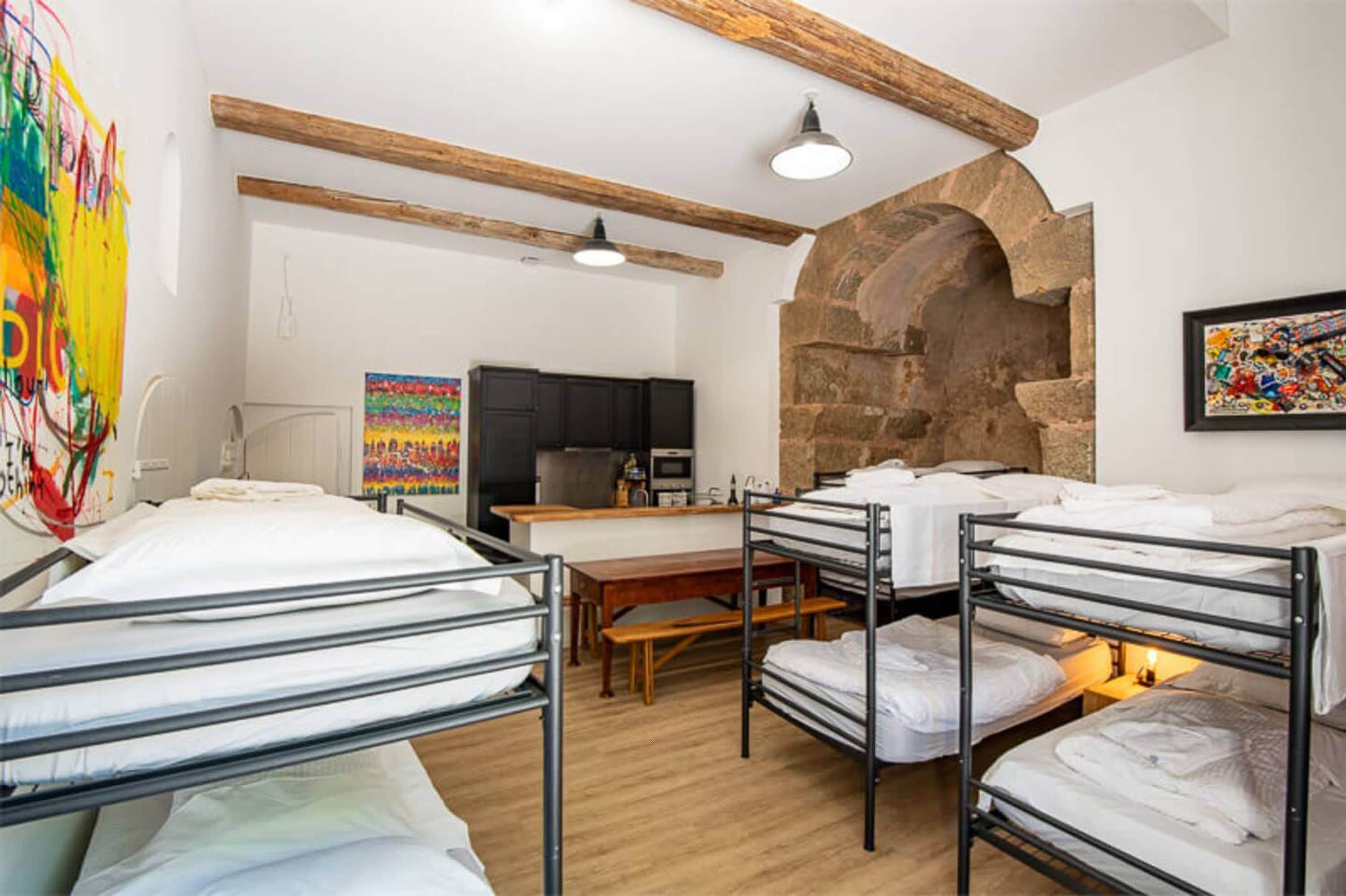 A dormitory room with bunk beds, a wooden table, a kitchen area, and a stone archway.