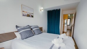 Un lit bien fait, avec une literie blanche et grise, une table de chevet, une lampe fixée au mur et une porte donnant sur une autre pièce.