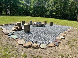 A circular fire pit surrounded by rocks and wooden posts, set in a grassy area with trees in the background. - Grandview Ridge, a Mountain Estate with scenic views and an unforgettable stay. (Schellsburg)