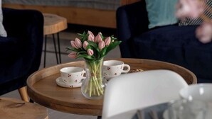 A cozy room with a bed, a round wooden table with a vase of flowers, and a dining setup with plates and cups. - Comfortable Apartment with Parking | Close to the Beach (Dziwnówek)