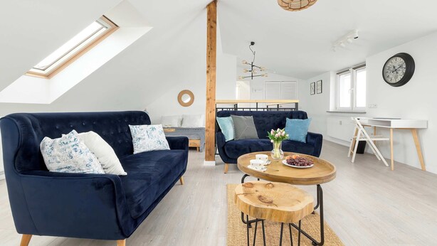 A modern loft apartment with a high ceiling, skylight, dark blue sofas, a wooden coffee table, and a white desk. - Comfortable Apartment with Parking | Close to the Beach (Dziwnówek)