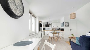 A modern kitchen with white cabinets, a dining area with a white table and chairs, and a blue sofa in the living area. - Comfortable Apartment with Parking | Close to the Beach (Dziwnówek)