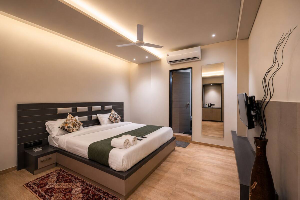 Un dormitorio moderno con cama, mesitas de noche, ventilador de techo, aire acondicionado, televisor y una pequeña kitchenette.