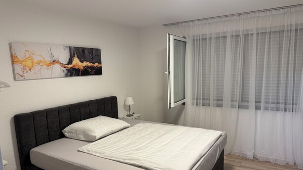 A modern bedroom with a bed, a nightstand, a lamp, a window with sheer curtains, and a wall art piece. - Modern Vacation Rental Near Phandtasialand: A Short Getaway 2 (Weilerswist)