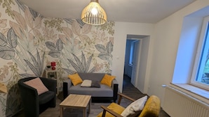 Ein Wohnzimmer mit Sofa, Sessel und Couchtisch, einer Wand mit Muster und einer Pendelleuchte, sowie einem Fenster mit Blick.