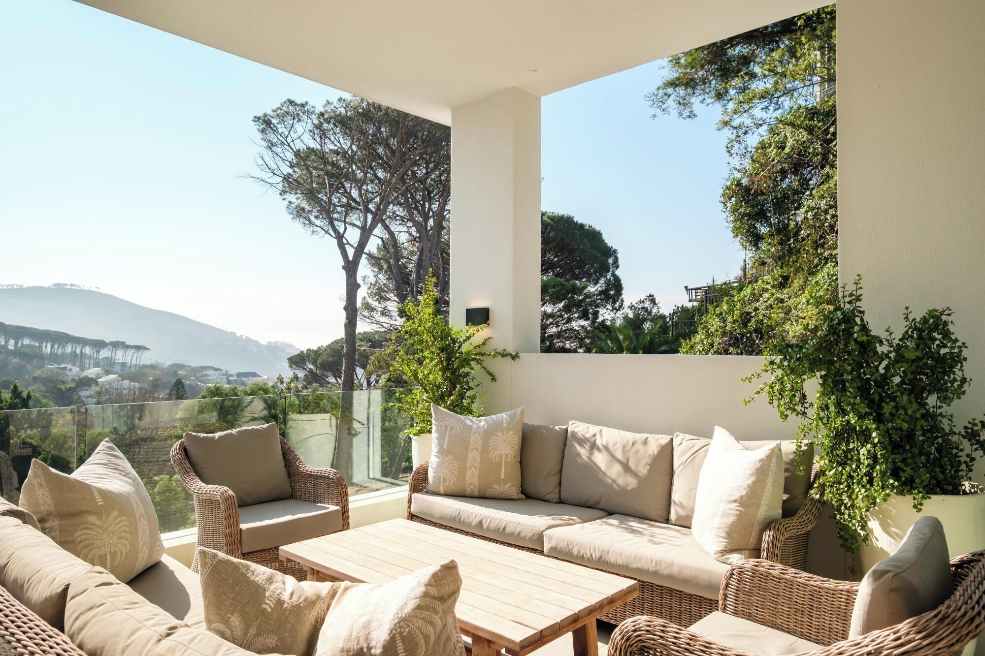 A sunlit outdoor seating area with wicker furniture, a wooden table, and a view of a hilly landscape.