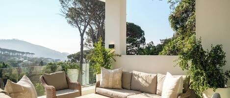 A sunlit outdoor seating area with wicker furniture, a wooden table, and a view of a hilly landscape.