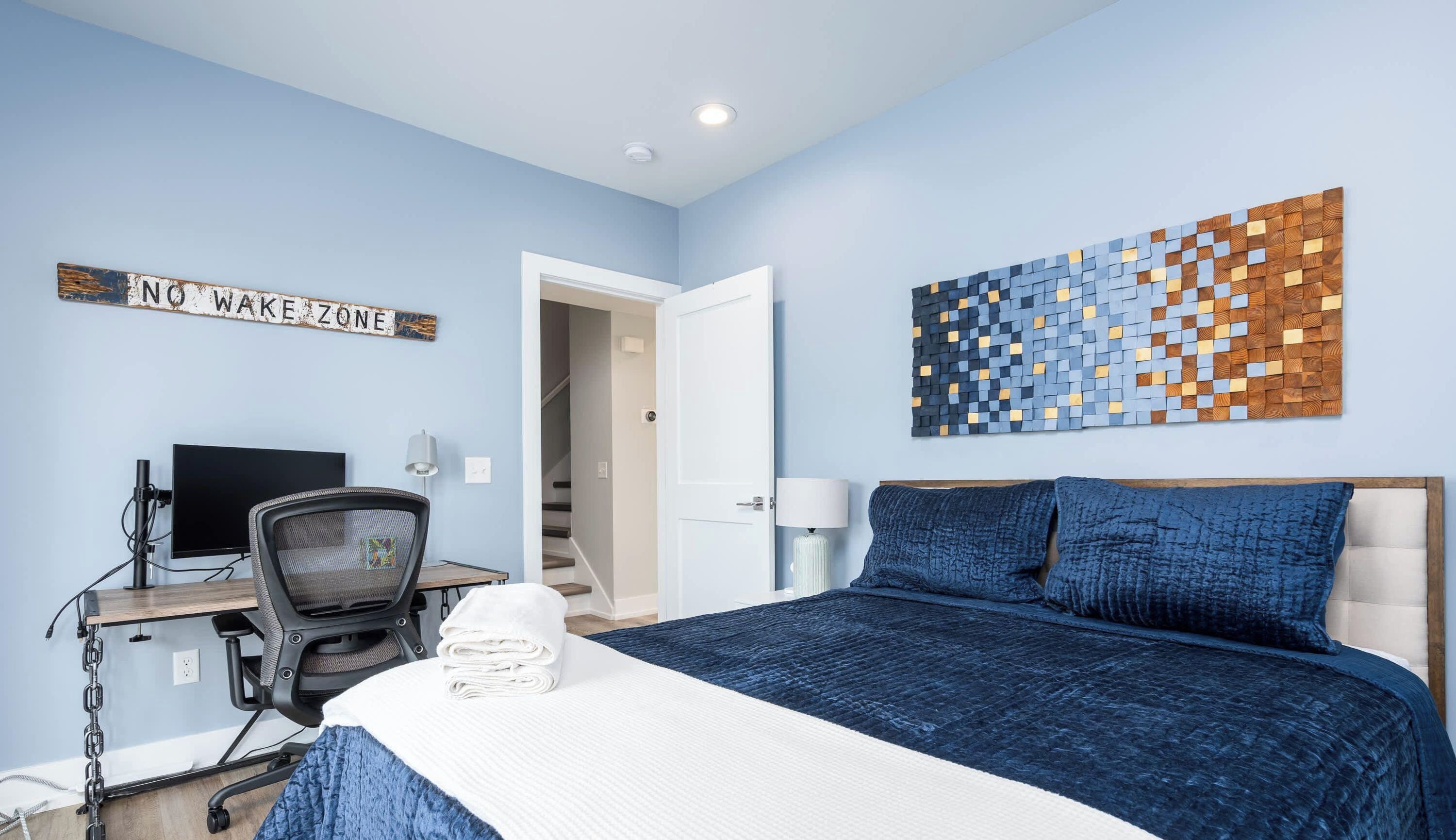 A bedroom with a bed, a desk, a chair, a wall art piece, and a sign that reads 'NO WAKE ZONE'.
